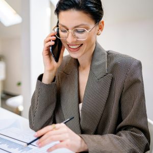 Confident businesswoman wearing glasses, talking on phone while reviewing documents in modern office setting.