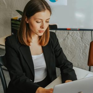 Confident businesswoman working on a laptop in a modern office environment with charts in the background.